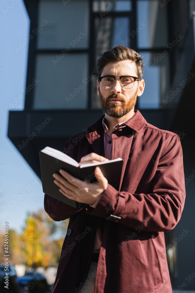Businessman in trench coat looking at camera while holding notebook outdoors. Businessman in trench coat looking at camera while holding notebook outdoors.