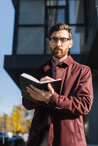 Businessman in trench coat looking at camera while holding notebook outdoors.