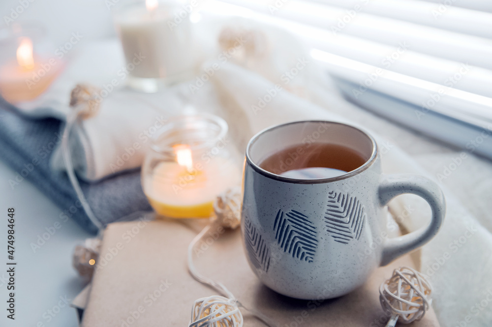 Cup of steaming tea, cozy mood. Burning candles and white sweaters on windowsill. Cozy autumn and winter concept, window with blinds...