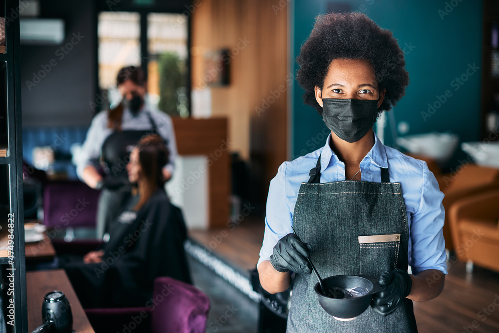 Fototapeta Black hairdresser wears face mask while working at hair salon during coronavirus pandemic.