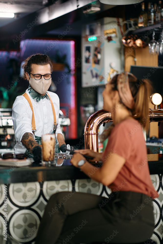 Happy bartender wears face mask while serving his customer at bar counter.