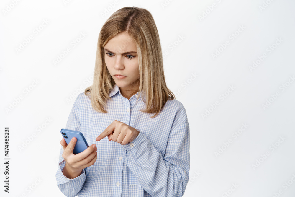 Image of blond little kid, girl staring at smartphone with shocked confused face, standing over white background Image of blond little kid, girl staring at smartphone with shocked confused face, standing over white background