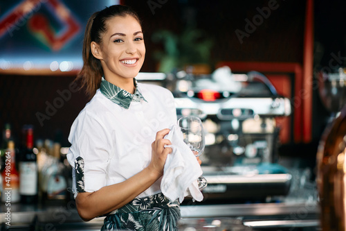 Wall Mural Happy female bartender cleans drinking glass at pub and looks at camera