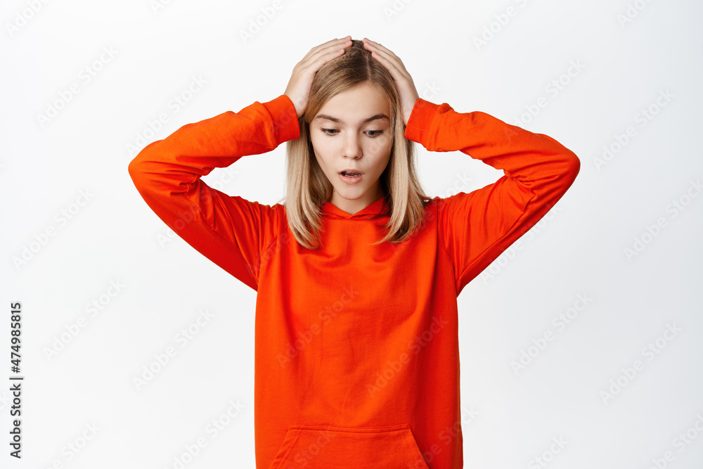Shocked and worried little girl, child in panic looking down, gasping startled, holding hands on head, standing overwhelmed against white background Shocked and worried little girl, child in panic looking down, gasping startled, holding hands on head, standing overwhelmed against white background