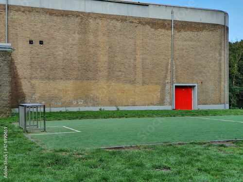 Photography Brick Wall with Bright Red Door and Green Grass Field with Soccer Goal in Amster