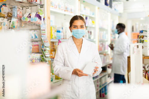 Portrait of a young female pharmacist in a protective mask, working in a pharmacy during the pandemic, standing 