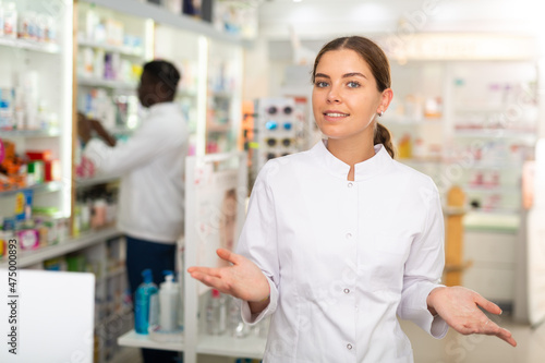 Portrait of female pharmacist standing in salesroom of drugstore and demonstrating haircare product. Her colleague working behind.