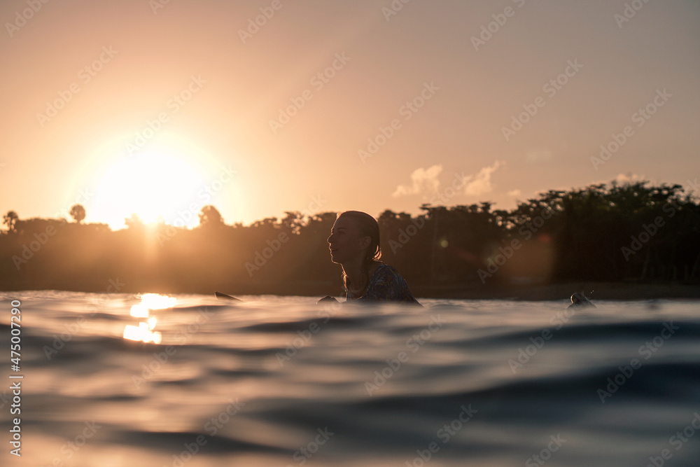 Portrait of blond surfer girl on white surf board in blue ocean pictured from the water at golden sunrise time in Encuentro beach