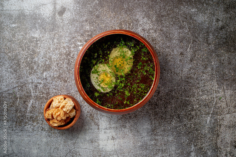 Chicken soup with boiled egg sprinkled with dill and parsley greens in a wooden bowl with bread crumbs stands isolated on a stone table, Flatlay Chicken soup with boiled egg sprinkled with dill and parsley greens in a wooden bowl with bread crumbs stands isolated on a stone table, Flatlay