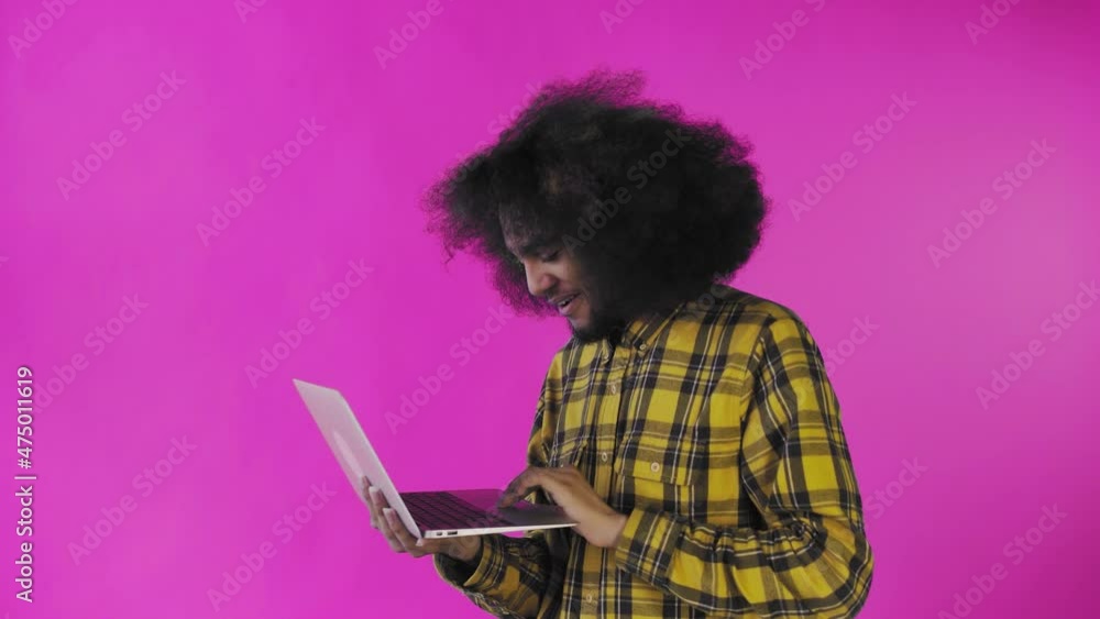 A young man with an African hairstyle on a pink background is typing on a laptop. On a colored background A young man with an African hairstyle on a pink background is typing on a laptop. On a colored background