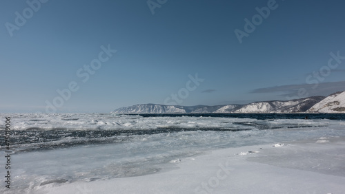 The ice on the river begins to melt. Footprints in the snow near the shore. In the distance - blue ice-free water. A mountain range against the azure sky. Siberia. Angara river