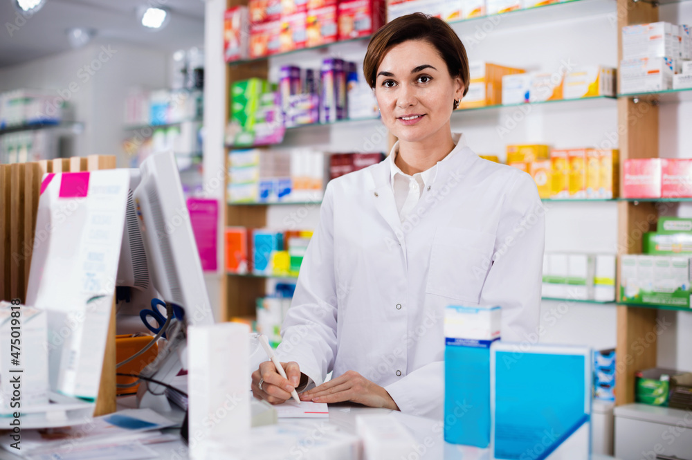 Young diligent smiling female pharmacist offering help in choosing at counter in pharmacy