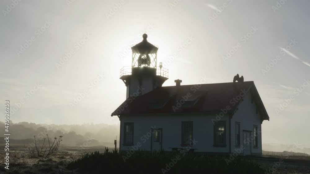 Sunrise scene on marine landmark in coastal park. Cinematic light house historic architecture building with rotating lantern and light beams illuminating foggy cloud with golden sun on background Sunrise scene on marine landmark in coastal park. Cinematic light house historic architecture building with rotating lantern and light beams illuminating foggy cloud with golden sun on background