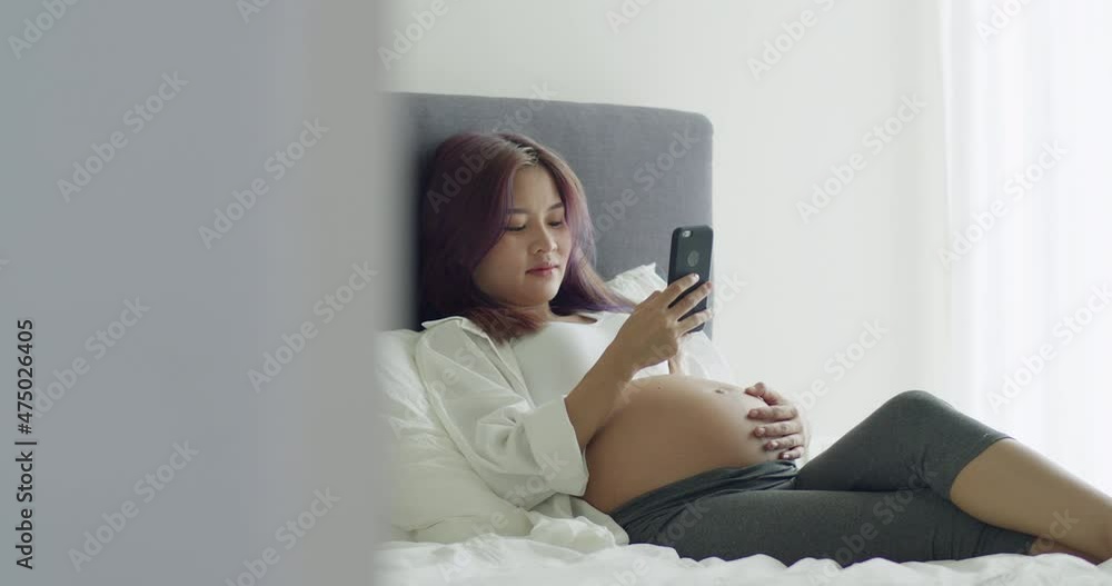 Cheerful young Asian pregnant wife using smartphone while lying on a bed in bedroom at home. Cheerful young Asian pregnant wife using smartphone while lying on a bed in bedroom at home.