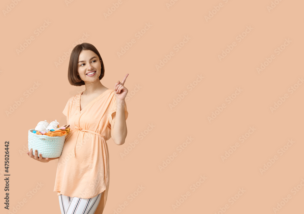 Young pregnant woman with baby clothes pointing at something on beige background Young pregnant woman with baby clothes pointing at something on beige background
