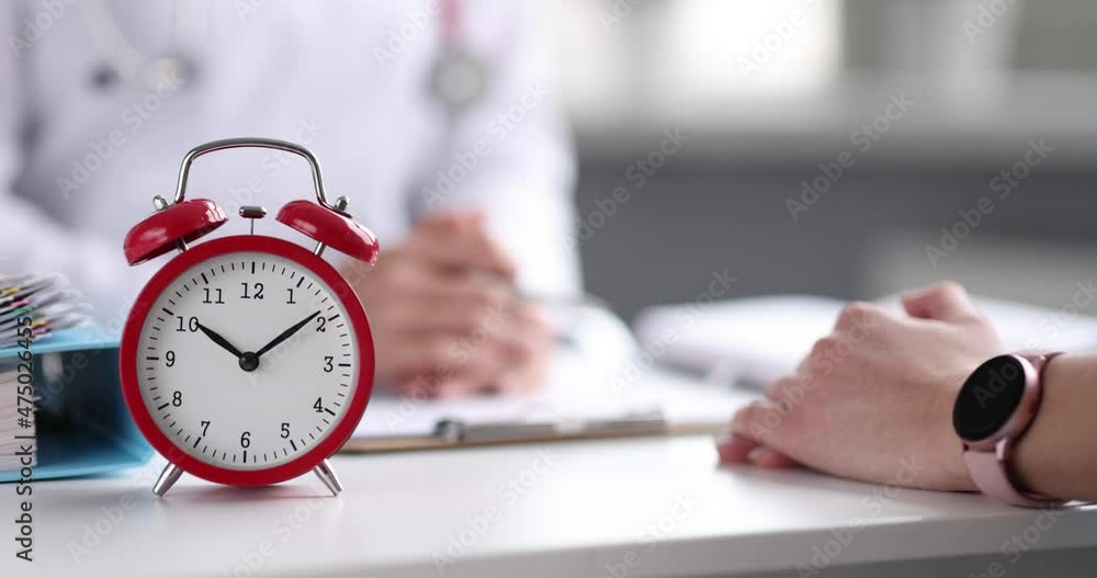 Doctor conducts appointment with patient in foreground with red alarm clock Doctor conducts appointment with patient in foreground with red alarm clock