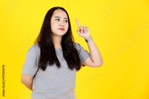 Portrait studio shot of Asian young chubby plump happy long black hair female model in gray casual tshirt outfit standing smiling pointing index finger up on blank copy space on yellow background