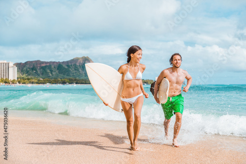 Hawaii surfing couple surfers in Waikiki beach, Honolulu Asian girl and Caucasian guy surfer carrying surfboards running out of water. Surf lifestyle Oahu island. USA travel. Fun vacation activity.