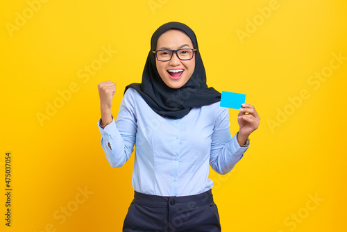 Portrait of excited young Asian woman holding a blank card and raising a fist in success gesture isolated on yellow background