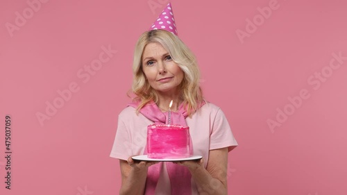 Sad distempered elderly blonde woman lady 40s years old wears t-shirt birthday hat look camera hold cake with candle sigh suspire sithe isolated on plain pastel light pink background studio portrait photo