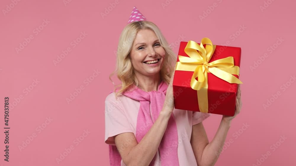 Smiling excited fancy elderly blonde woman lady 40s years old wears t-shirt birthday hat hold shake red present box with gift ribbon bow isolated on plain pastel light pink background studio portrait Smiling excited fancy elderly blonde woman lady 40s years old wears t-shirt birthday hat hold shake red present box with gift ribbon bow isolated on plain pastel light pink background studio portrait