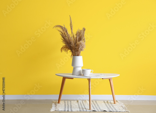 Obraz na plátně Vase with dry reeds, cup and newspaper on table near yellow wall