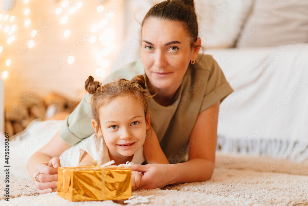 mom and funny daughter in white dress sits on floor on rug with gift in gold wrapping paper. tradition to give gifts to children for New Year and birthday. Santa Claus. children's toy store