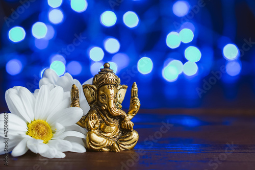 Golden lord ganesha sculpture in daisy flowers over blue illuminated background. Copy space for text