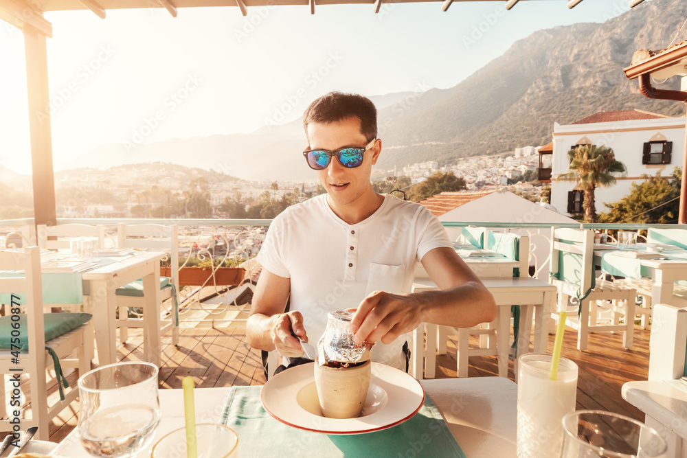 Young man eating Testi kebab in restaurant on a terrace. It is an authentic Turkish delicious meat dish baked in a clay pot, served after breaking the jug. - obrazy, fototapety, plakaty 