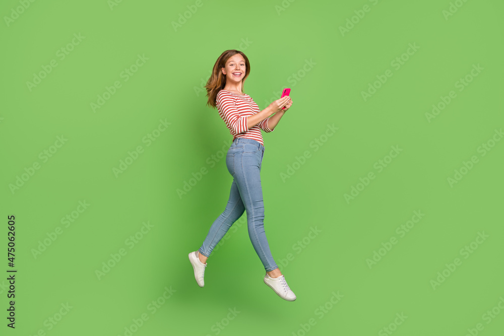 Photo of carefree influencer lady jump hold phone wear striped shirt jeans footwear isolated green color background Photo of carefree influencer lady jump hold phone wear striped shirt jeans footwear isolated green color background