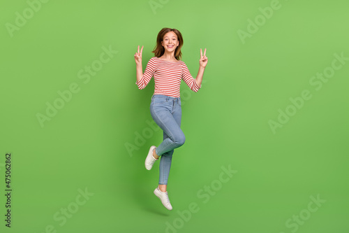 Photo of cheerful friendly lady jump show v-signs wear striped shirt jeans sneakers isolated green color background