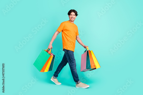 Full body profile photo of optimistic brown hair young guy go with bags wear t-shirt jeans sneakers isolated on blue background