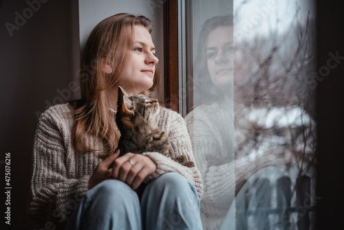 Young cheerful girl sitting at home on the windowsill in a warm sweater playing with a gray cat on a winter day photo