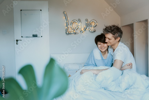 Loving couple in pajamas having fun in bed in the bedroom with word Love on the wall background. They are embracing and laughing. Happy family mornings at home. Valentine's day. Selective focus. © okrasiuk