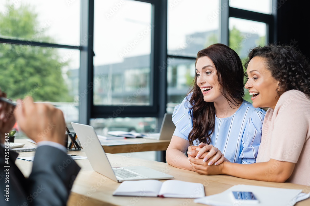 Fototapeta premium joyful interracial lesbians holding hands while looking at laptop near blurred realtor