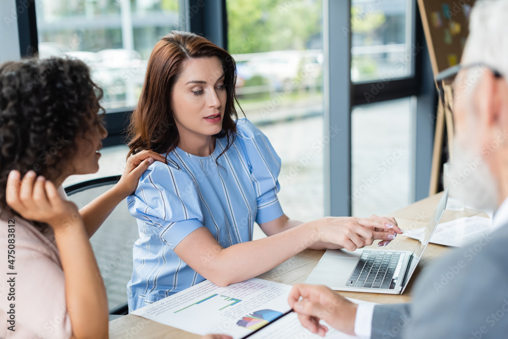 Obraz premium african american woman touching shoulder of lesbian girlfriend pointing with finger at laptop near blurred realtor