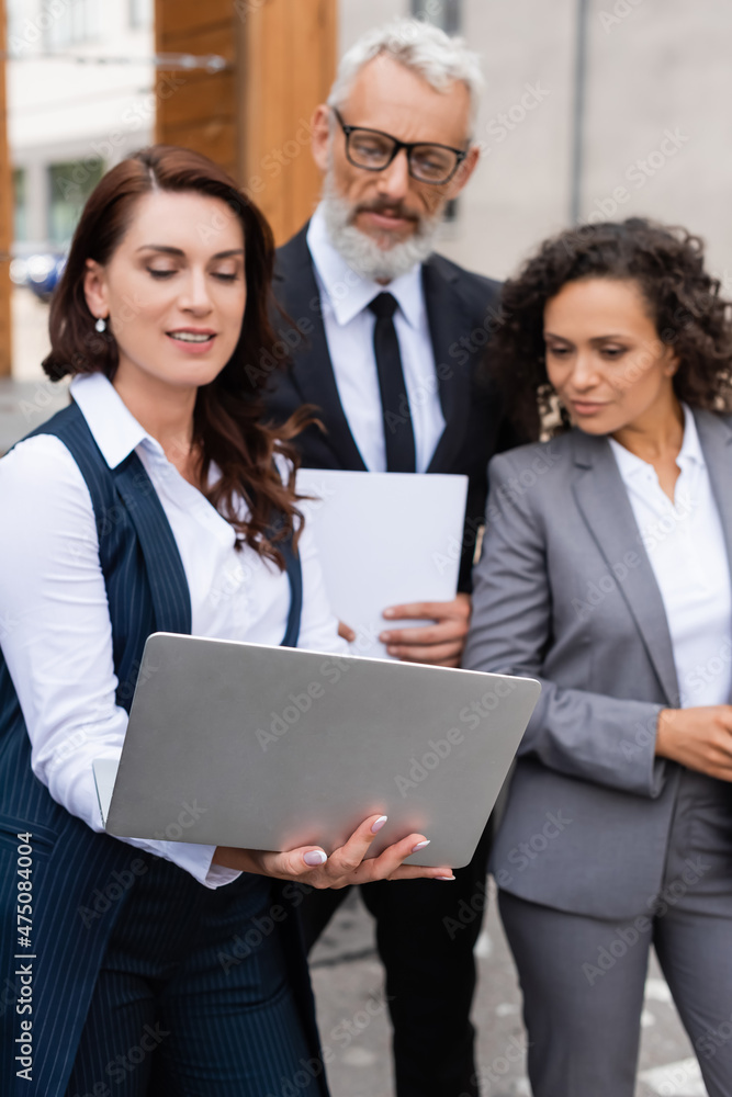 real estate broker holding laptop near african american woman and blurred middle aged businessman real estate broker holding laptop near african american woman and blurred middle aged businessman