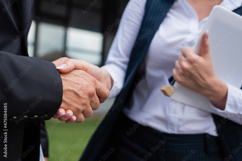 cropped view of blurred real estate agent with key and digital tablet shaking hands with businessman outdoors