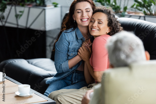 happy multiethnic lesbian women with closed eyes holding hands while sitting on couch near blurred psychologist