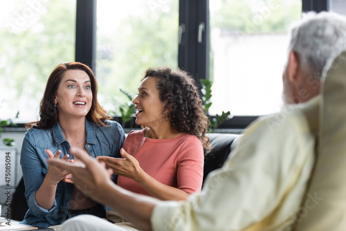 excited multiethnic lesbian couple gesturing while talking with blurred psychologist during appointment