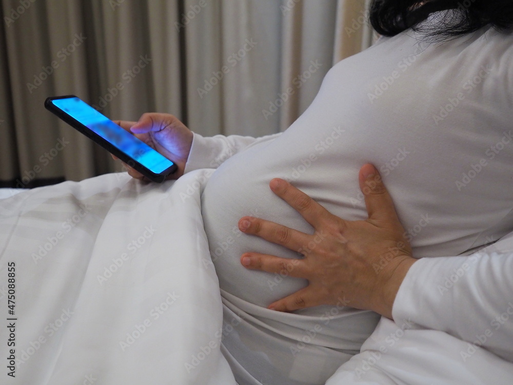 Pregnant Asian woman wearing a white t-shirt. The white blanket was stroking his stomach as he sat on the bed while studying the pregnancy information from his mobile phone with interest.
