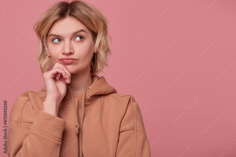 Studio portrait of young blonde woman with curly hair, wears beige hoodie thoughtfully looks aside at copy space. touching her chin, isolated over pink background Studio portrait of young blonde woman with curly hair, wears beige hoodie thoughtfully looks aside at copy space. touching her chin, isolated over pink background