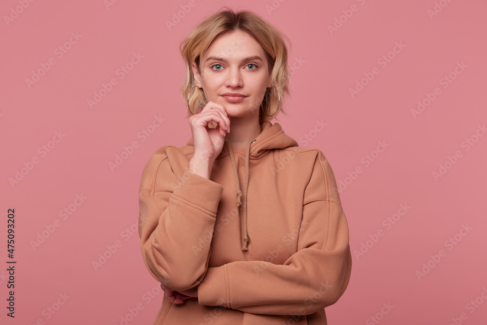 Studio portrait of young blonde woman with curly hair, wears beige hoodie thoughtfully looks into camera and smiles. touching her chin, isolated over pink background Studio portrait of young blonde woman with curly hair, wears beige hoodie thoughtfully looks into camera and smiles. touching her chin, isolated over pink background