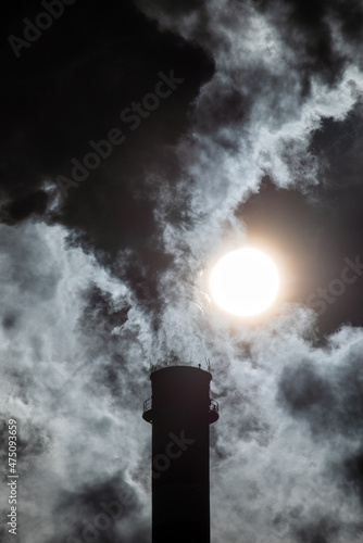 heavy smoke coming out of a huge and high factory chimney against the sun on the sky