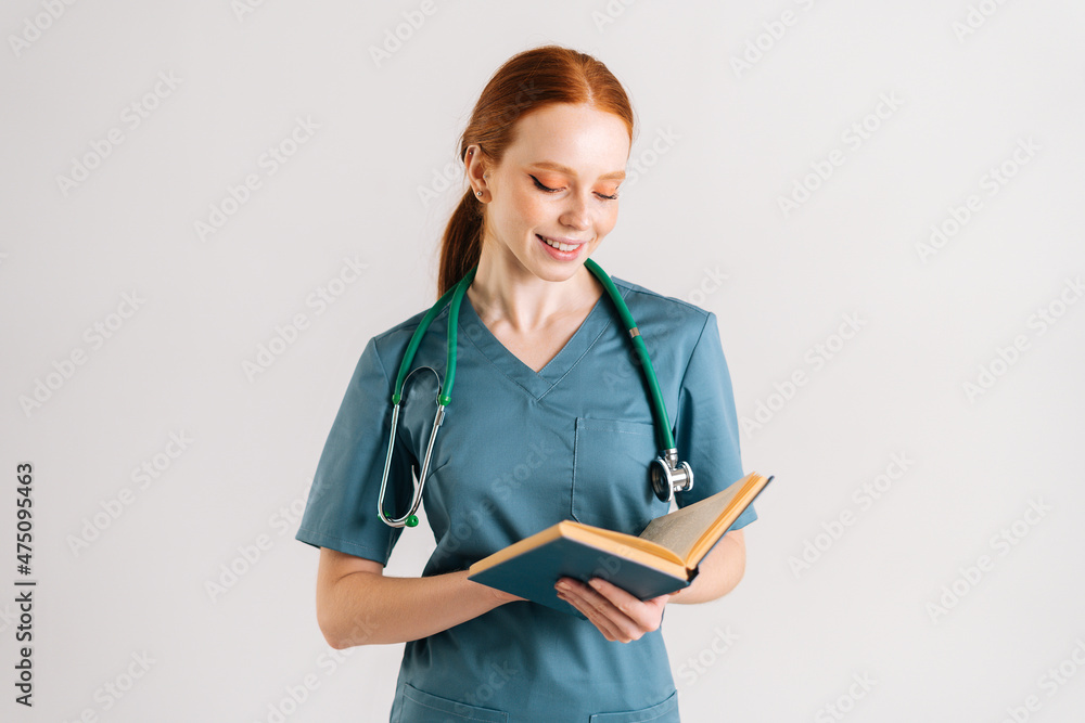 Portrait of positive young woman physician in green uniform with stethoscope reading medical book standing on white isolated background in studio. Female doctor reading paper treatise. Portrait of positive young woman physician in green uniform with stethoscope reading medical book standing on white isolated background in studio. Female doctor reading paper treatise.