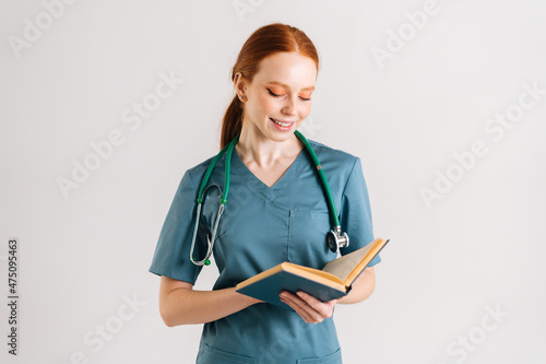 Portrait of positive young woman physician in green uniform with stethoscope reading medical book standing on white isolated background in studio. Female doctor reading paper treatise.