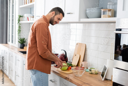 Side view of smiling man looking at digital tablet while cutting vegetables in kitchen