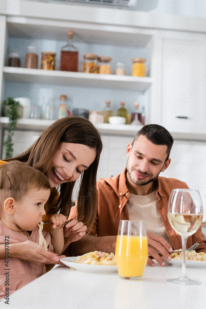 Woman feeding son with pasta near drinks and husband at home