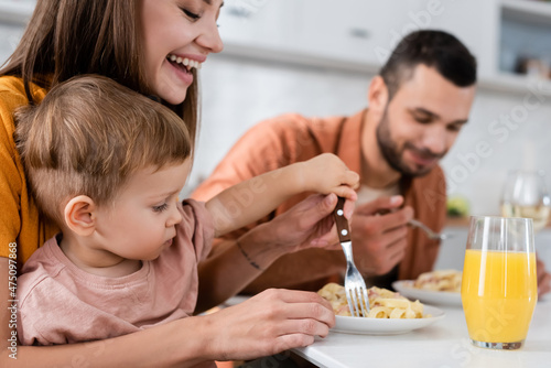 Happy woman and son holding fork near pasta and orange juice in kitchen
