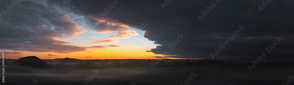Panorama of a dramatic colorful sunset with dark and bright clouds against the backdrop of mountains. Mountains and forest in fog on the background of sunset with gloomy clouds.